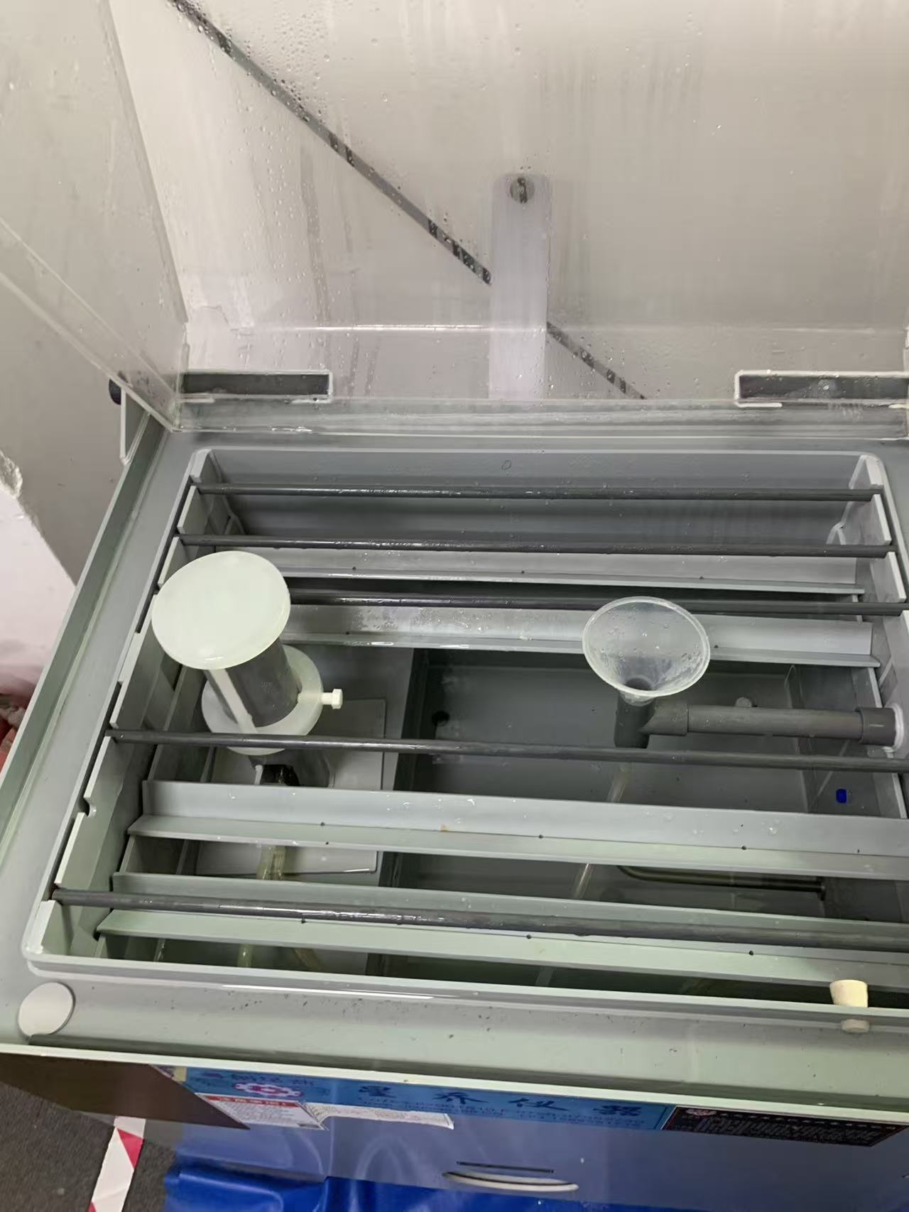 Interior of the salt spray chamber showing the atomising tower, glass collection funnel with graduated cylinder, and specimen supports